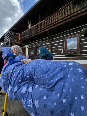 Eine ältere Frau sitzt unter einer blauen Decke mit Sternenmustern, während sie auf eine andere Person blickt, die sich in der Nähe befindet. Im Hintergrund ist ein Holzhaus mit einem Balkon zu sehen, das einige Schäden aufweist.