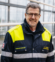Ein lächelnder Mann in Arbeitskleidung mit einer gelb-reflektierenden Jacke steht vor einer modernen, gläsernen Struktur. Er trägt eine Brille und hat einen kurzen Bart. Im Hintergrund sind strahlende Fenster zu sehen.