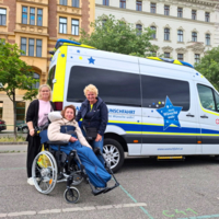 Drei Personen, eine im Rollstuhl, stehen vor einem Wunschfahrzeug.