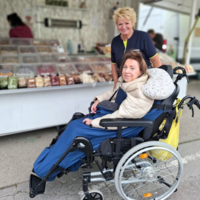 Eine Frau im Rollstuhl lächelt, während eine Betreuerin hinter ihr steht.