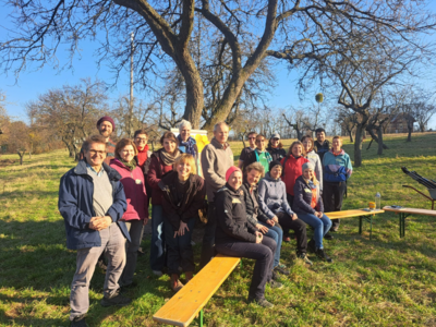 Gruppe von Menschen in einem Obstgarten, versammelt auf Bänken im Freien.