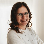 Frau mit Brille und lächelndem Gesicht in einem hellen Raum.