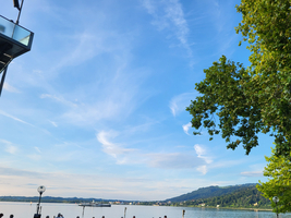 Menschen stehen an einem Gewässer unter einem blauen Himmel mit Wolken.