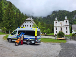 Ein Rettungswagen steht auf einem Parkplatz vor einer Kirche und einem Haus, umgeben von grünen Bäumen und Bergen. Zwei Personen, eine im Rettungsdienst-Outfit und eine andere mit einem Rollstuhl, unterhalten sich. Es ist bewölkt und die Umgebung wirkt ruhig und ländlich.