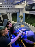 Drei Personen in einem Krankenwagen bieten einer älteren Person, die auf einer Liege liegt, Becher an. Der Raum ist schwach beleuchtet und wirkt freundlich, mit einer Decke und Spielzeug im Hintergrund. Es zeigt eine herzliche Interaktion und Fürsorge.