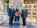 Drei Personen stehen zusammen, eine in Uniform mit Blumenstrauß und Urkunde.