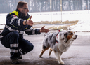 Ein Mann in einer Sicherheitsuniform kniet auf dem Boden und winkt einem Australian Shepherd, der auf ihn zusteuert. Der Hintergrund zeigt Schnee und Bäume, was auf eine kalte Jahreszeit hinweist.