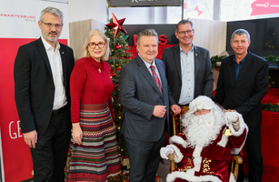 Fünf Personen stehen um einen Weihnachtsmann vor einem festlich geschmückten Baum.