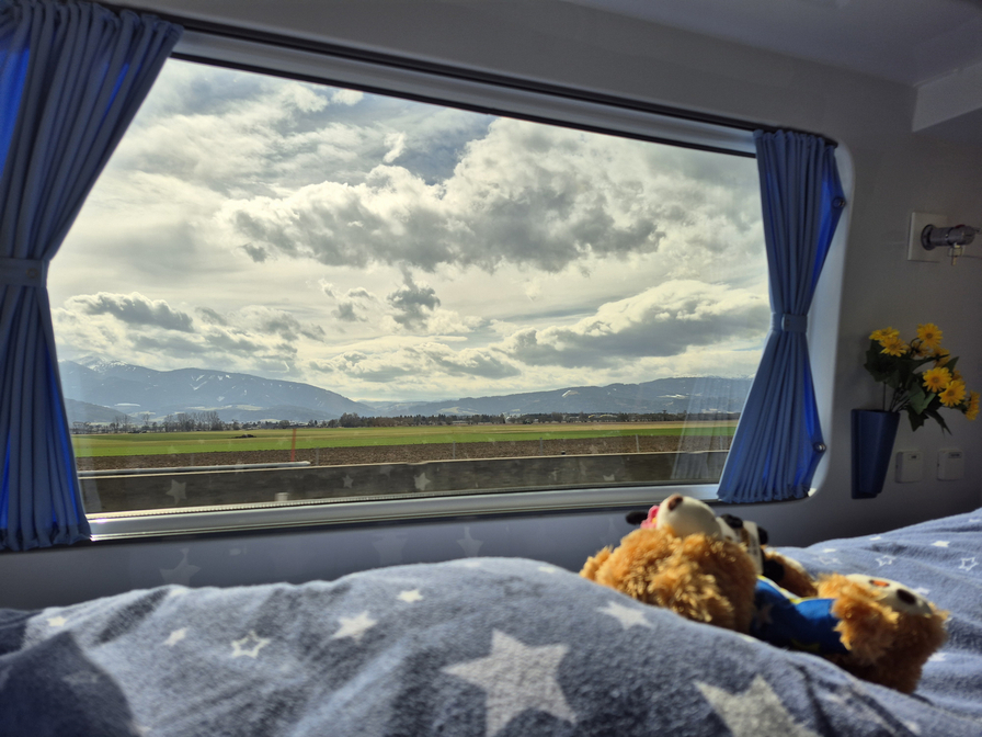 Blick aus dem Fenster eines Zuges, das eine landschaftliche Szene mit Bergen und Wolken zeigt. Im Vordergrund ist ein Bett mit einer Decke, auf der ein Teddybär liegt. Ein Blumenstrauß steht im Fenster.